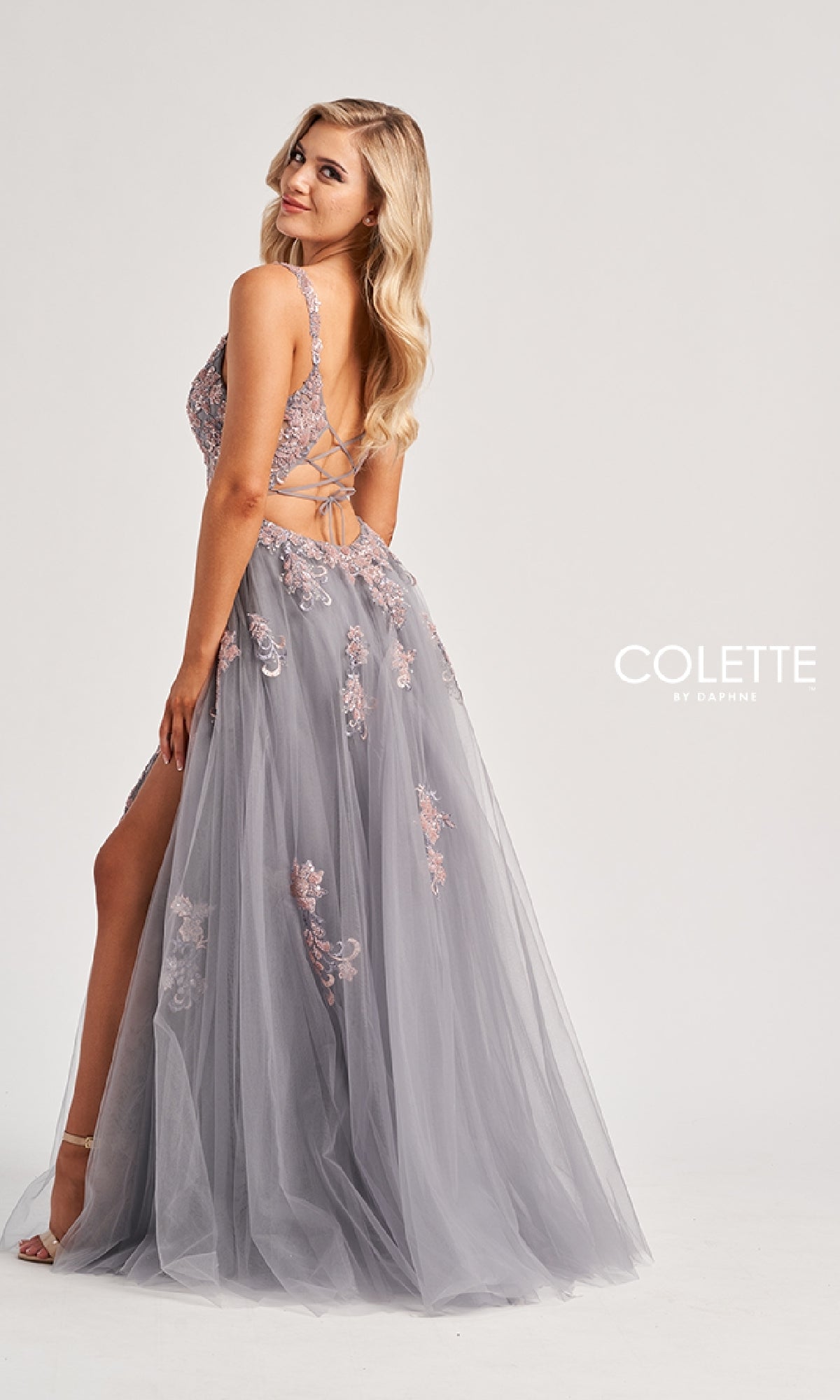 A woman models the Colette CL8130, a lavender long prom dress featuring a tulle skirt, lace-up strappy open back, delicate floral embroidery, lace embellishments, and a thigh-high slit against a plain light background.